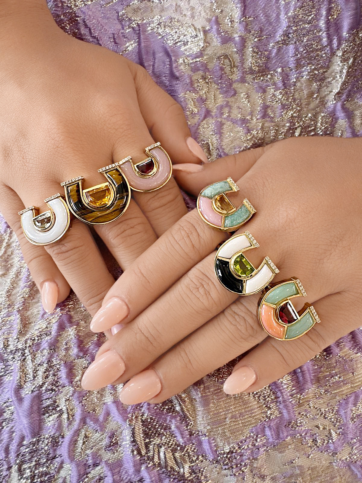 A close-up view of two hands modeling a variety of Sig Ward Jewelry 14K yellow gold horseshoe rings inlaid with colorful gemstones, enamel, and pave diamonds from the Manifest Collection.