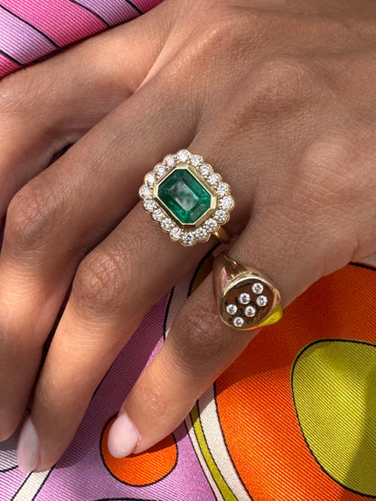 Close-up of a hand wearing two rings with a green emerald with a diamond halos and a diamond pinky ring on a colorful Pucci background.
